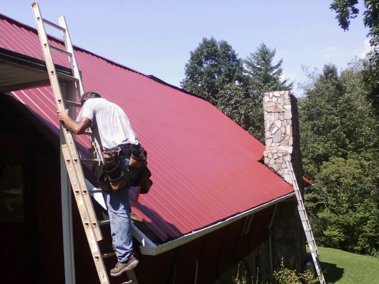Roofer on a ladder beside a red metal roof and stone chimney in West Alexander